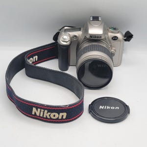 Peut inclure: Un appareil photo reflex argentique Nikon F55 argenté avec un objectif noir, une poignée noire et une sangle noire portant le logo Nikon en blanc avec des bordures rouges. Un bouchon d'objectif circulaire noir est posé à côté du boîtier.
