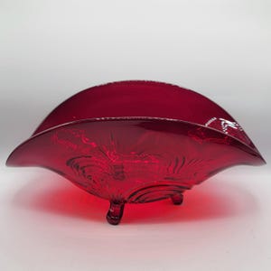 Könnte beinhalten: Eine rubinrote Glasschale mit ausgestelltem, gewelltem Rand und drei kleinen Füßen. Die Schale hat ein strukturiertes, wirbelndes Muster im Inneren und ein zartes weißes Design entlang des Randes. Ein dekoratives Stück zum Servieren oder Ausstellen.