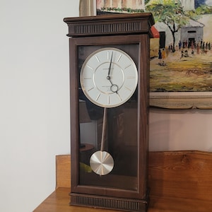 May include: A rectangular wall clock with a dark brown wooden frame and a glass front panel. The clock face is circular with a silver finish, featuring thin black hour markers and black hands. A silver pendulum hangs inside the wooden case.