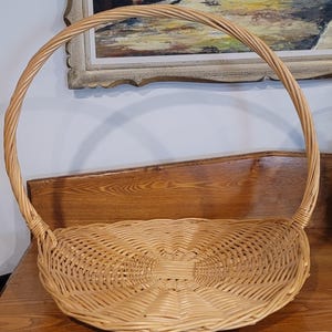 Op de afbeelding: Een lichtbruine rieten mand met een grote, gebogen handgreep. De mand heeft een ronde, ondiepe bodem en is gemaakt van geweven riet. De handgreep is aan de zijkanten van de mand bevestigd. De mand staat op een houten oppervlak.