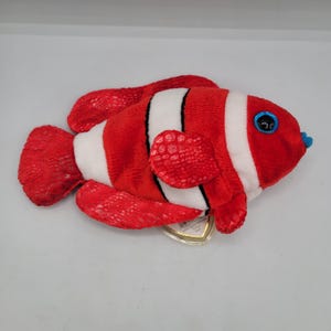 May include: A plush toy clownfish in red, white, and black. The fish has large, round blue eyes and a small blue mouth. The fins and tail have a textured pattern. A small tag is attached.