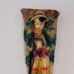 May include: A vintage wall pocket vase featuring a painted figure of a person in traditional attire. The figure wears a hat, jacket, and shorts, holding a staff. The vase has a textured background with foliage and is primarily beige, green, and brown.