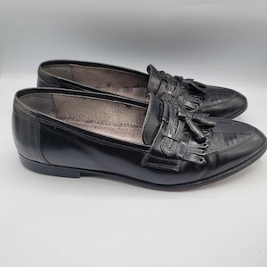May include: A pair of black leather loafers with tassel details. The shoes have a classic design with a rounded toe and a low heel. The loafers are displayed on a white background.