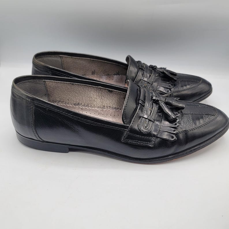 May include: A pair of black leather loafers with tassel details. The shoes have a classic design with a rounded toe and a low heel. The loafers are displayed on a white background.