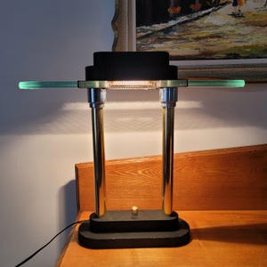 Puede incluir: Lámpara de escritorio de estilo Art Déco con base y pantalla negras. La lámpara tiene dos soportes metálicos dorados y un elemento horizontal de vidrio verde claro. La lámpara está encendida, emitiendo un brillo cálido.