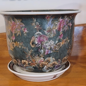 May include: A round ceramic planter with a dark teal, mottled background featuring a detailed floral and bird pattern in shades of pink, white, and gold. The planter has a wide, flared white rim and sits on a matching white ceramic saucer.