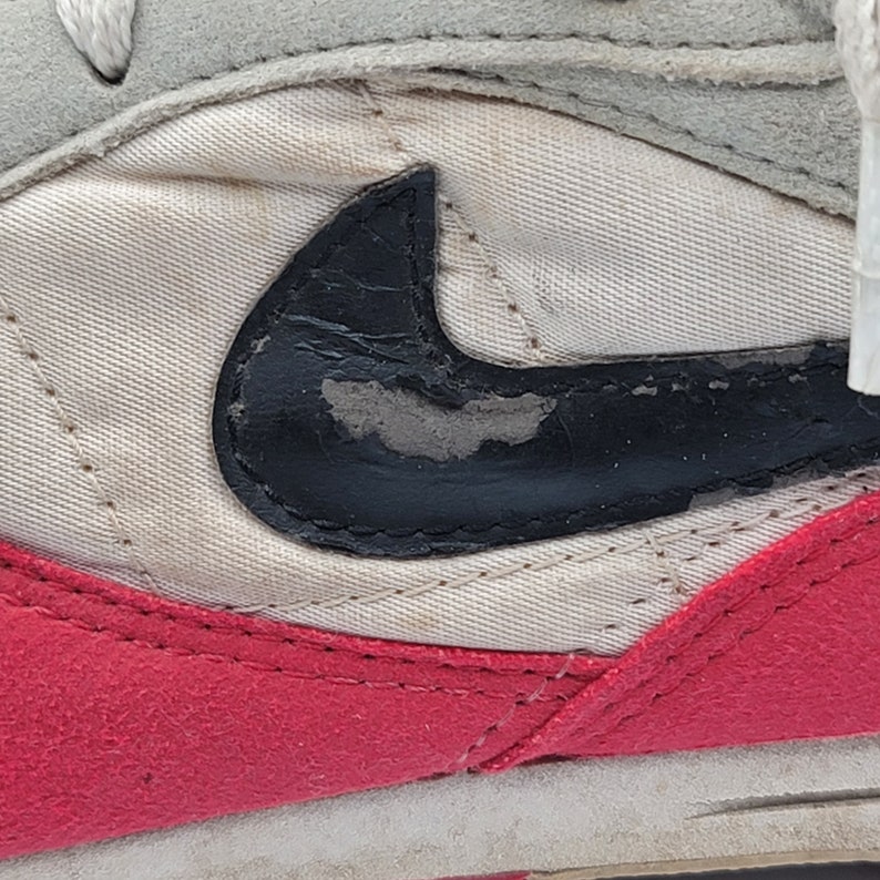 May include: Close-up of a white and red sneaker with a black Nike swoosh logo. The swoosh is made of a textured material and has a worn look.