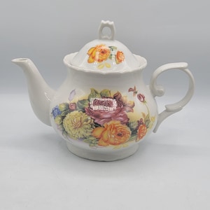 Puede incluir: Tetera de cerámica blanca con tapa y asa. La tetera está decorada con un estampado floral con rosas en tonos amarillos, naranjas y rosas. La tapa tiene un pequeño asa de bucle.