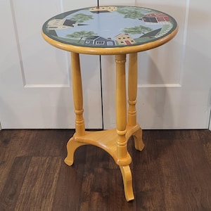 May include: A round wooden side table with a light natural finish, featuring three turned legs and a triangular base. The tabletop is painted with a blue sky background, green trees, and several small houses and a red barn in a circular pattern.
