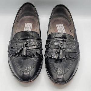 May include: A pair of black leather loafers with a textured, reptile-like pattern on the toe and vamp. Each shoe features a decorative strap with a tassel and fringe detail. The brand name "CALDERO" is visible on the inner sole.