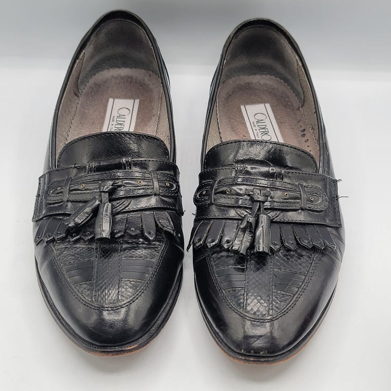 May include: A pair of black leather loafers with a textured, reptile-like pattern on the toe and vamp. Each shoe features a decorative strap with a tassel and fringe detail. The brand name "CALDERO" is visible on the inner sole.