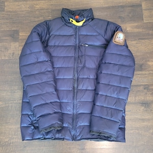 May include: A dark navy blue quilted puffer jacket featuring a full-length front zipper, a high collar with a yellow strap detail, and a brown circular patch on the left sleeve. The jacket has horizontal stitching throughout and a chest pocket.