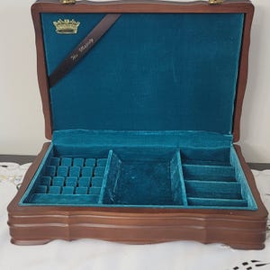 May include: Open wooden jewelry box with a teal velvet interior. The box has multiple compartments for storage, including slots for rings and a larger space for other items. A gold crown emblem and the words "His Majesty" are visible.