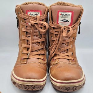 May include: A pair of brown leather lace-up boots with side zippers and faux fur lining. The tongue features a red and white Pajar brand patch with mountain graphics and text. The boots have a thick rubber sole with a red stripe detail.