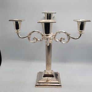 Puede incluir: Un candelabro de metal plateado con un pilar central de estrías verticales, una base cuadrada y cuatro brazos que se extienden hacia afuera. Cada brazo y la columna central sostienen un portavelas de parte superior cuadrada con acabado liso.