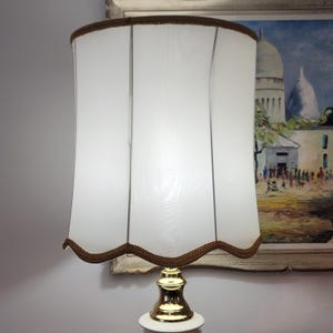 May include: A table lamp featuring a cylindrical white fabric shade with vertical panels and a scalloped bottom edge trimmed in brown fabric. The lamp base is made of polished gold-tone metal with a smooth, reflective finish and a white circular stand.