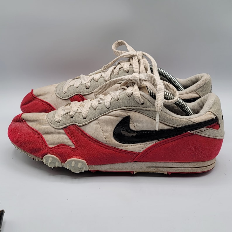 May include: White and red running shoes with a black Nike swoosh logo. The shoes have a white sole with small spikes.