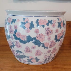 Peut inclure: Un grand cache-pot rond en céramique blanche orné d'un motif peint à la main de fleurs roses, de feuilles bleues et de petits fruits. Le récipient présente un bord large et évasé ainsi qu'une finition lisse et brillante avec des motifs floraux.