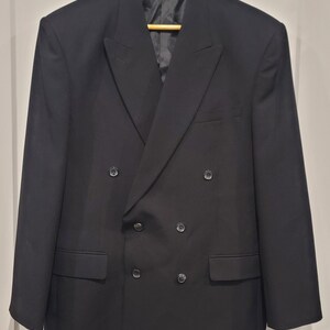 May include: A black double-breasted blazer with six buttons and two front pockets. The blazer is hanging on a wooden hanger. The jacket has a notched lapel and a tailored fit. The fabric appears to be a smooth, solid color.