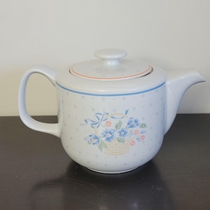 Peut inclure: Théière en céramique blanche avec couvercle, anse et bec verseur. La théière est ornée d'un motif de panier floral bleu et d'un ruban bleu. Le couvercle a une bordure orange et un petit bouton.