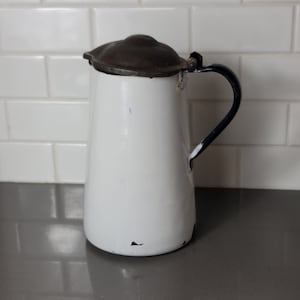 Könnte beinhalten: Weißer Emaille-Krug mit dunkelblauem Henkel und braunem Metalldeckel. Der Krug hat eine leicht abgeplatzte Emaille-Oberfläche.