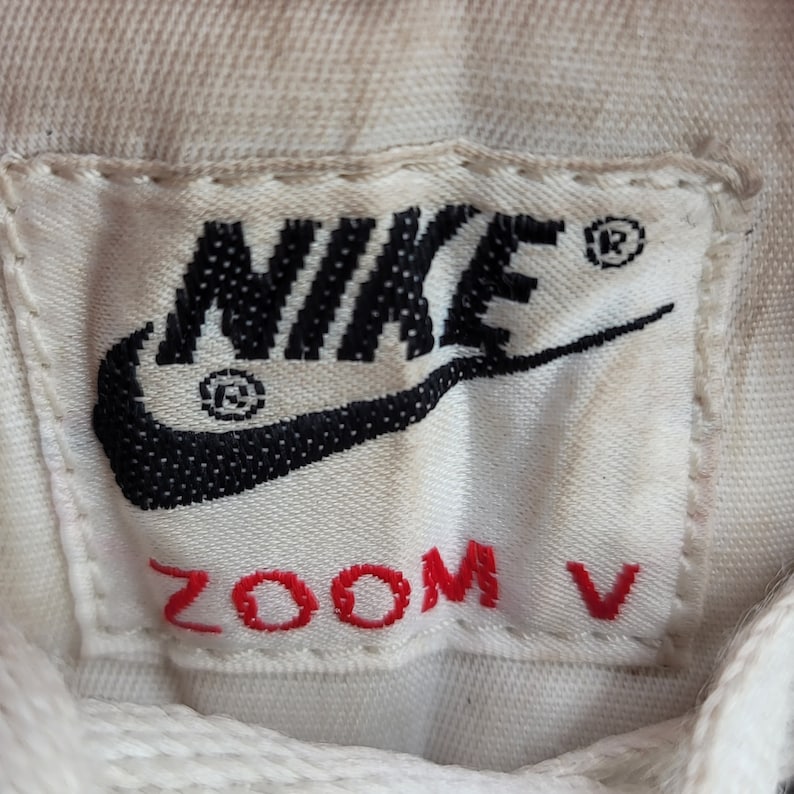 May include: White athletic shoe with a black Nike swoosh logo and red "ZOOM V" text on the tongue.