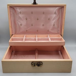 May include: Open vintage jewelry box with a light beige exterior and a pink interior. The interior features multiple compartments and a decorative pattern on the lining. The box has a metal clasp and a keyhole.