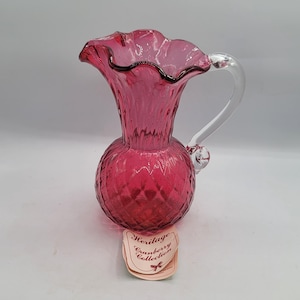 Puede incluir: Una jarra de cristal color rojo arándano con borde ondulado y cuerpo con patrón de diamantes. La jarra tiene un asa de cristal transparente y una etiqueta en la base que dice Heritage Cranberry Collection con un pequeño lazo rojo.