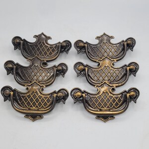 Puede incluir: Un juego de seis tiradores de cajón de metal en tono latón antiguo, cada uno con un asa decorativa con patrón de celosía y bordes curvos ornamentados. Los herrajes tienen un acabado oscuro y desgastado con reflejos en las superficies.