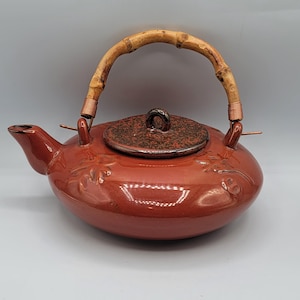 May include: A red ceramic teapot with a bamboo handle and a brown lid. The teapot has a floral design.