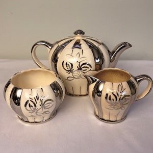 May include: A white teapot, creamer and sugar bowl with silver accents and floral designs. The set is made of ceramic and has a vintage style.