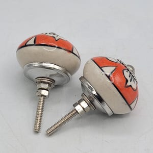 May include: Two round ceramic cabinet knobs featuring a cream-coloured base with a vibrant orange and black geometric floral pattern. Each knob includes a silver-toned metal base, a threaded bolt, and a nut for secure installation on furniture.