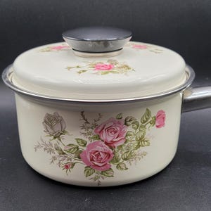 Könnte beinhalten: Cremefarbener Emaille-Kochtopf mit Deckel, verziert mit rosa und grauen Rosenblütenmustern. Der Topf hat einen silbernen Rand und Griff. Der Deckel hat einen schwarzen Knopf. Ideal zum Kochen.