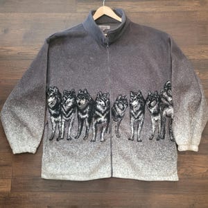 Peut inclure: Veste polaire grise zippée avec un motif de loup répété sur la partie inférieure. Les loups sont représentés en noir et blanc. La veste a un col montant et une fermeture éclair intégrale. Les manches ont un effet dégradé.