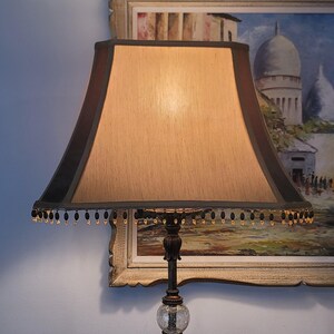 May include: A table lamp featuring a beige, square-tapered fabric shade with dark brown trim and a dangling beaded fringe. The lamp base includes a dark bronze-toned metal stem and a clear, textured glass sphere at the bottom of the stand.