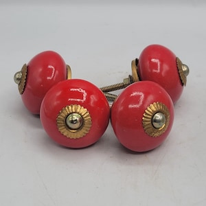 May include: A set of four round, glossy red ceramic cabinet knobs. Each knob features a central metallic silver-toned screw head surrounded by a decorative, scalloped brass-colored metal plate. The knobs include threaded metal bolts for mounting.