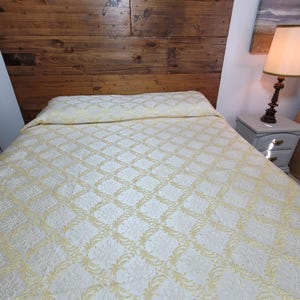 May include: A bedspread featuring a cream and gold diamond pattern with intricate floral motifs within each section. The fabric has a subtle sheen and covers the entire bed surface, including a matching pillow sham at the head of the bed.