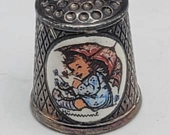 May include: A vintage silver thimble with a detailed design. The thimble features a painted oval with a child in a red hood and blue clothing. The thimble has a textured, diamond-patterned side and a dimpled top.