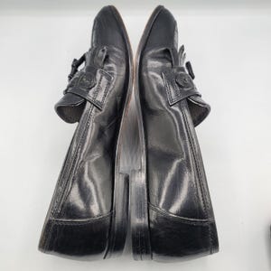May include: Pair of black leather loafers with decorative fringe and buckle details. The shoes have a classic design with a polished finish, suitable for formal or business attire. The soles are a contrasting brown color.