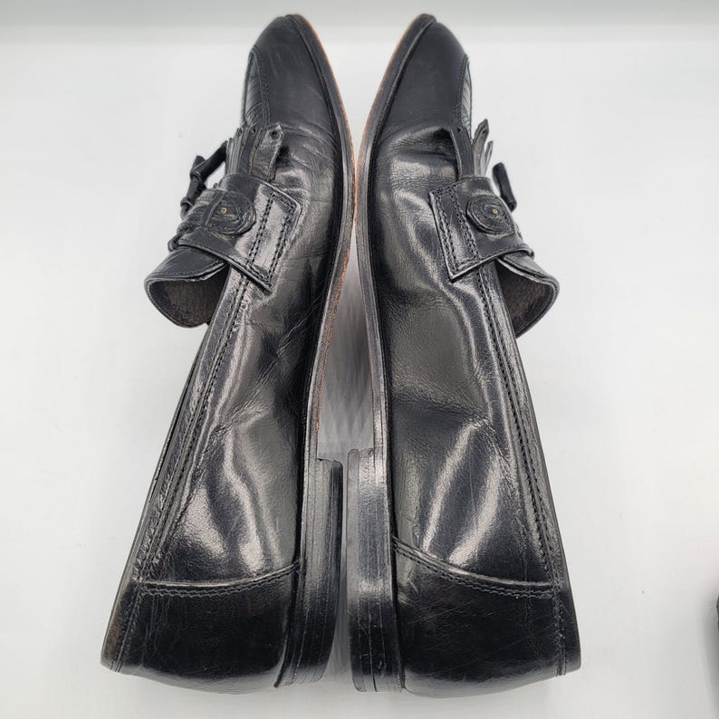 May include: Pair of black leather loafers with decorative fringe and buckle details. The shoes have a classic design with a polished finish, suitable for formal or business attire. The soles are a contrasting brown color.