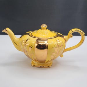 Puede incluir: Una tetera de cerámica de color amarillo brillante con un acabado lustroso y detalles en oro metálico. La tetera tiene un asa curva, un pico puntiagudo y una tapa con un pequeño pomo. Está decorada con patrones florales dorados.