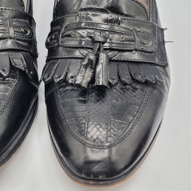 May include: Close-up of a pair of black leather loafers. The shoes feature a textured vamp, decorative tassels, and fringe detailing. The leather appears to be in good condition, with a polished finish. The shoes have a classic, timeless design.