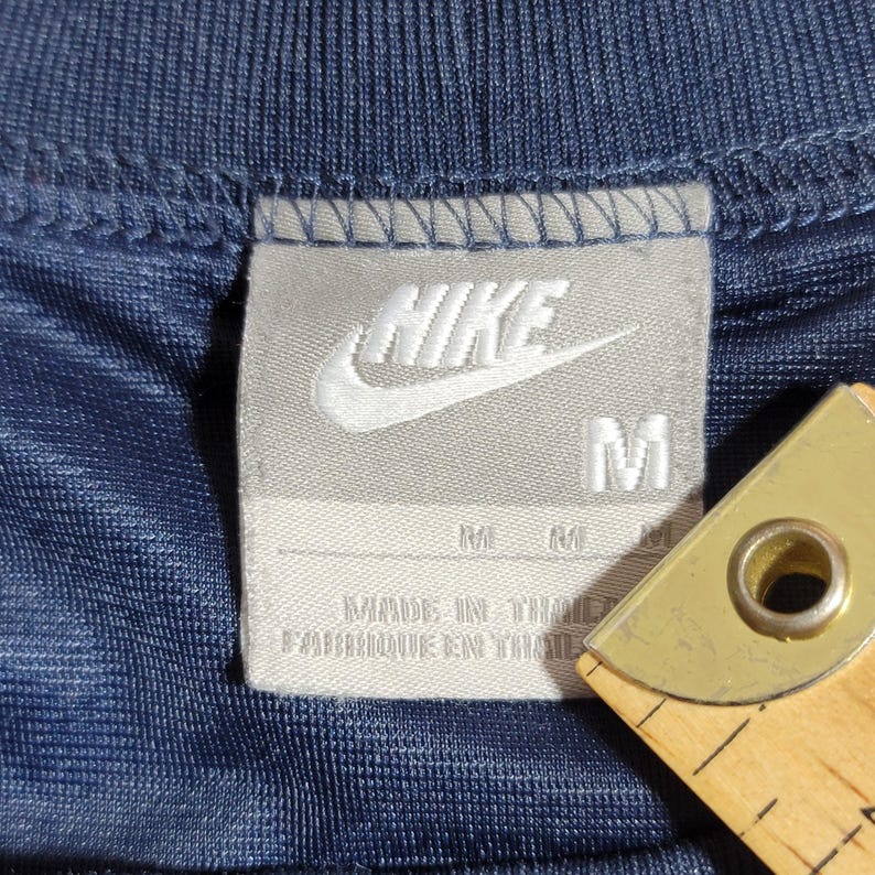 Puede incluir: Una camiseta azul marino con un logotipo de Nike blanco y una etiqueta blanca que dice "Nike" y "M" para el tama&ntilde;o. La etiqueta tambi&eacute;n dice "Made in Thailand" y "Fabriqu&eacute; en Tha&iuml;lande".