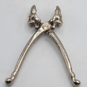 Puede incluir: Un cascanueces de metal plateado con dos diseños de cabezas de animales esculpidas en el punto de pivote superior. La herramienta tiene dos mangos largos y curvos que se estrechan hacia extremos redondeados, unidos por un mecanismo de bisagra central.