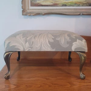 May include: A rectangular footstool featuring a light gray and cream damask fabric upholstery. The piece is supported by four curved, metallic legs with a bronze-toned finish, each ending in a rounded foot design, providing a stable base.
