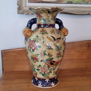 May include: A large, cream-coloured ceramic vase with a flared top and two dark blue handles. The vase is adorned with colourful floral and bird motifs. The base and rim feature a dark blue geometric pattern. A decorative piece for home decor.