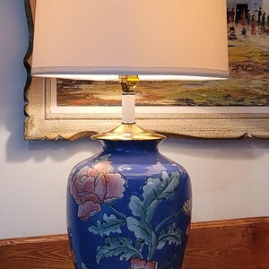 May include: A blue ceramic table lamp with a beige lampshade. The lamp base features a floral design in pink and green. The lamp is approximately 71 cm tall.