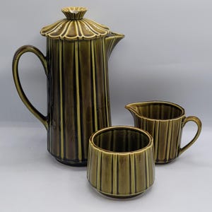 May include: A vintage green ceramic coffee set comprising a tall coffee pot with a lid, a creamer, and a sugar bowl. The set has vertical ribbed detailing and a glossy finish. The coffee pot is approximately 25 cm tall.