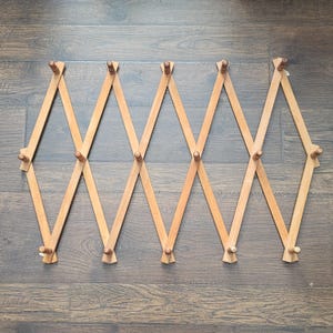 May include: A light-coloured wooden expandable coat rack with multiple pegs. The rack is in a diamond-shaped design, with pegs evenly spaced along the wooden arms. The rack is shown against a dark wood grain background.
