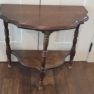 Puede incluir: Una mesa consola de madera de color marrón oscuro en forma de media luna con un tablero semicircular de borde festoneado. La mesa se apoya en tres patas torneadas e incluye un estante inferior semicircular con acabado de veta.